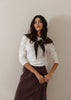 A woman modeling a white long-sleeve top with a dark brown knit collar featuring white trim, paired with a dark brown skirt, against a plain beige wall.