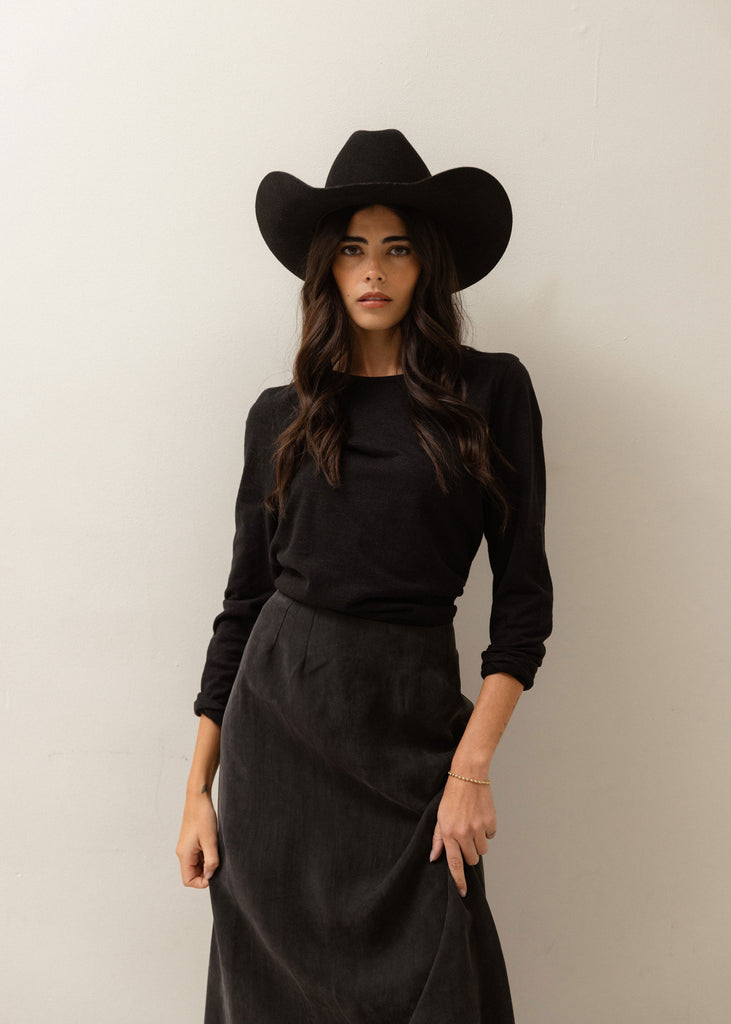 A woman wearing a black wide-brim cowboy hat, a black long-sleeve top, and a black midi skirt, standing against a plain light gray wall.