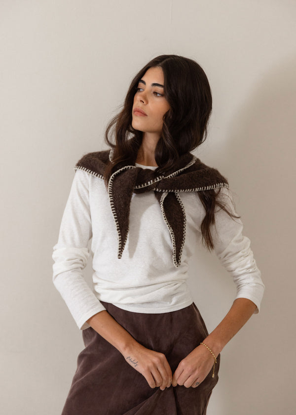 A woman wearing a white long-sleeve top and a dark brown skirt, featuring a prominent dark brown knit collar with white trim, posed against a plain light beige wall.