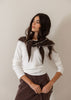 A woman wearing a white long-sleeve top and a dark brown skirt, featuring a prominent dark brown knit collar with white trim, posed against a plain light beige wall.