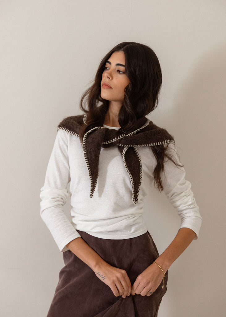 A woman wearing a white long-sleeve top and a dark brown skirt, featuring a prominent dark brown knit collar with white trim, posed against a plain light beige wall.