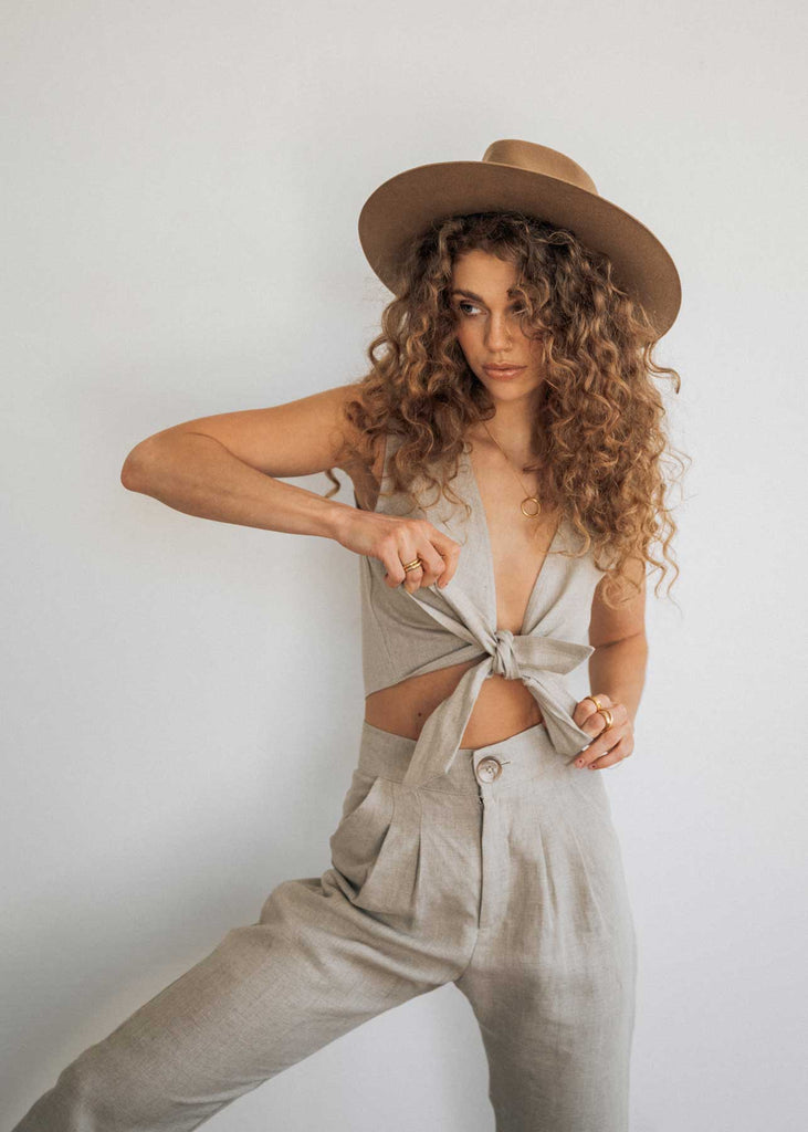 The Ballina Tie Up and Trouser in Oat. Made from 100% linen in Vancouver, BC. Our model is wearing a hat over her curly hair and holding the ties of the top while standing against a blank white wall.