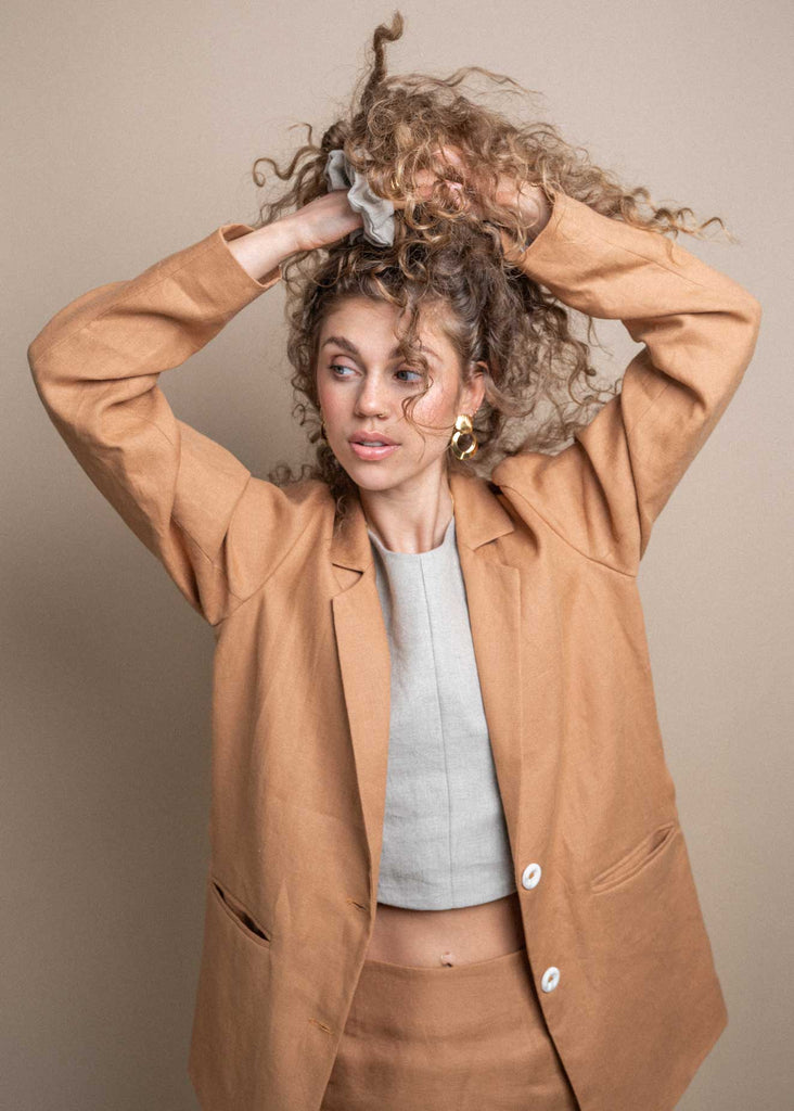 The Sienna Blazer in Camel paired with our Slate Scrunchie and shot in studio. Our gorgeous model shows off our linen clothes while tying her curly locks.